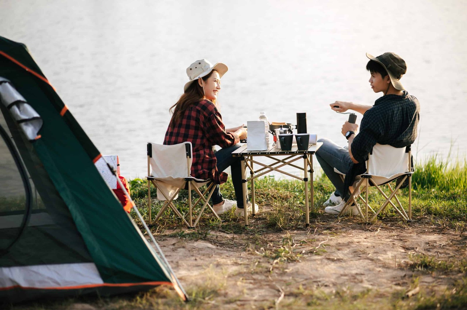 froehliches junges backpacker paar das vor dem zelt im wald mit kaffeesatz sitzt und beim campingausflug im sommerurlaub frische kaffeemuehle macht scaled